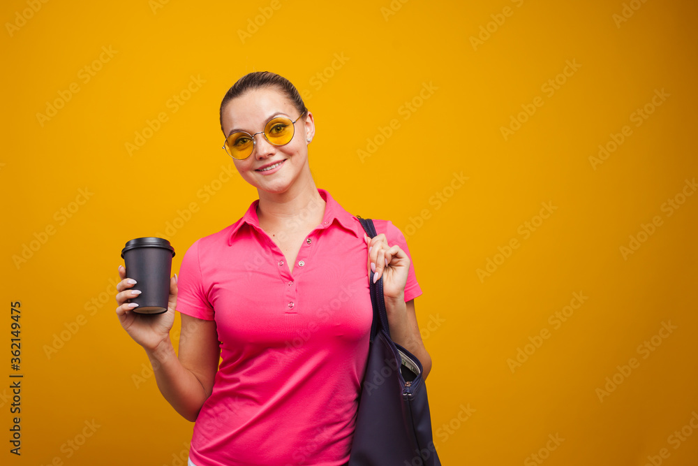 Cute blonde in sunglasses and t-shirt with bag on shoulder, drinking her hot drink, portrait on yellow background Cute blonde in sunglasses and t-shirt with bag on shoulder, drinking her hot drink, portrait on yellow background
