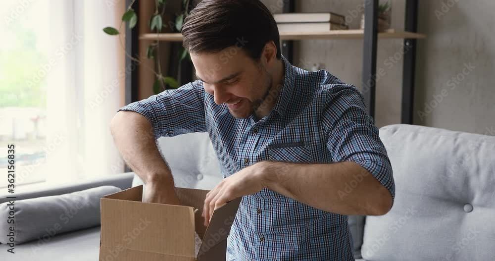 Millennial guy sitting on sofa at home in cozy living room opening delivered parcel small cardboard box feels glad. Good qualified, easy quick delivery services satisfied client. Distant order concept Millennial guy sitting on sofa at home in cozy living room opening delivered parcel small cardboard box feels glad. Good qualified, easy quick delivery services satisfied client. Distant order concept
