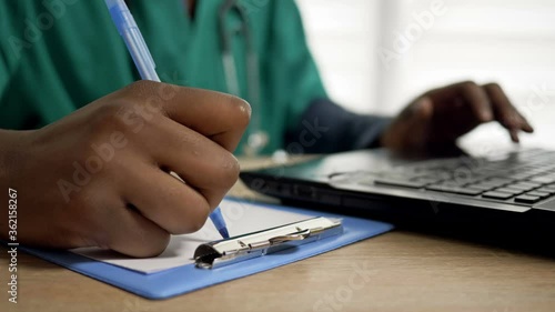 Black doctor works at a computer and makes notes in his office. Close up of doctor hands.