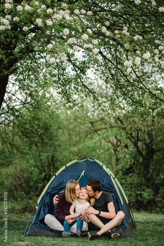 Mother, father kisses and hugs a child enjoying a camping holiday in the countryside. Happy family holidays in a tent with a child in nature. Сoncept of summer vacation and travel, trip. Campsite.