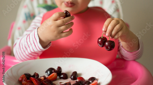 Baby girl eating cherries and strawberries with one hand and holding  two cherries with the another hand