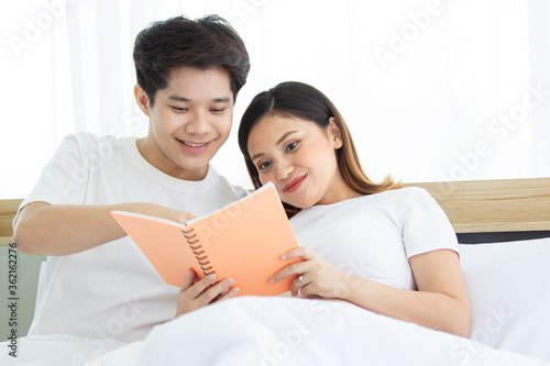 Portrait of lovely husband and wife reading a book together on the bed in bedroom, husband embracing his pregnant wife on a bed during reading a book. Couple relaxing together. Family leisure activity