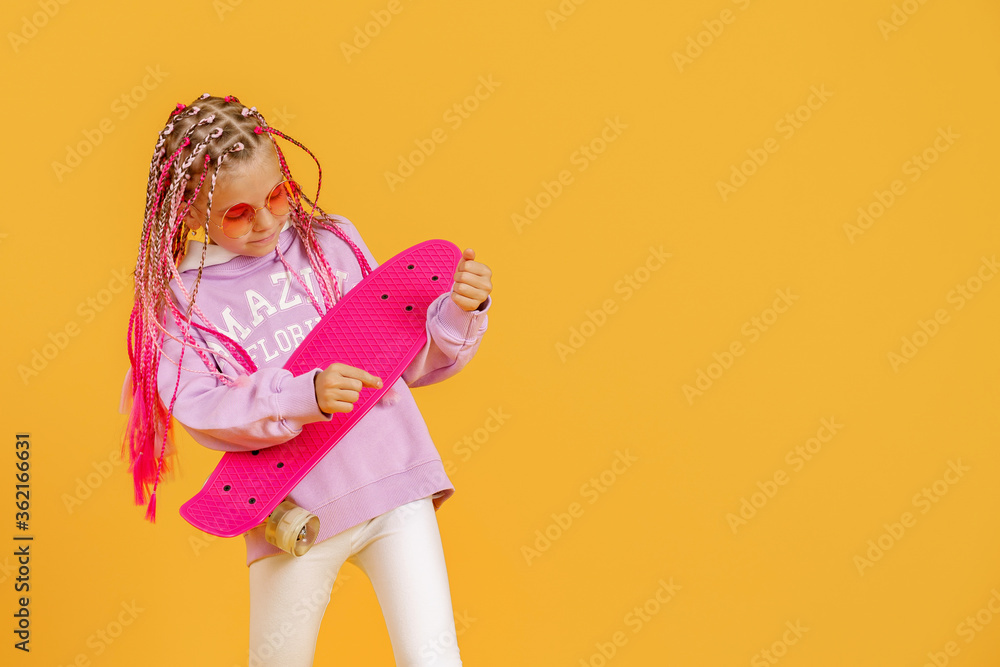 Active young girl in pink shirt and white shorts holding skateboard over yellow background.
