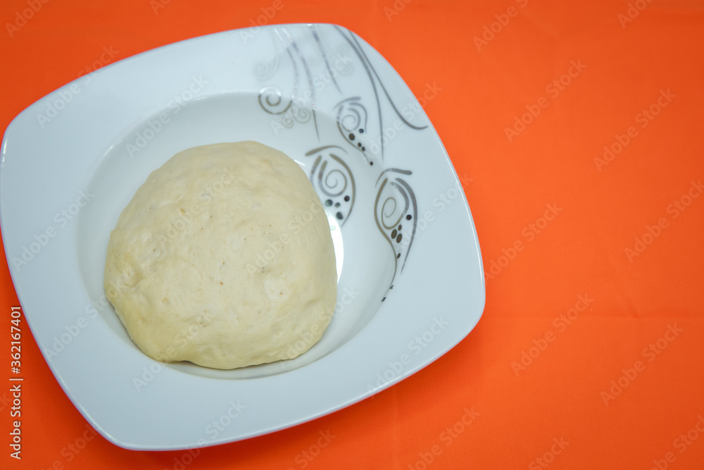 Raw dough lying on the plate on a white background. A piece of dough for bread baking . The dough is on a white plate on an orange background .