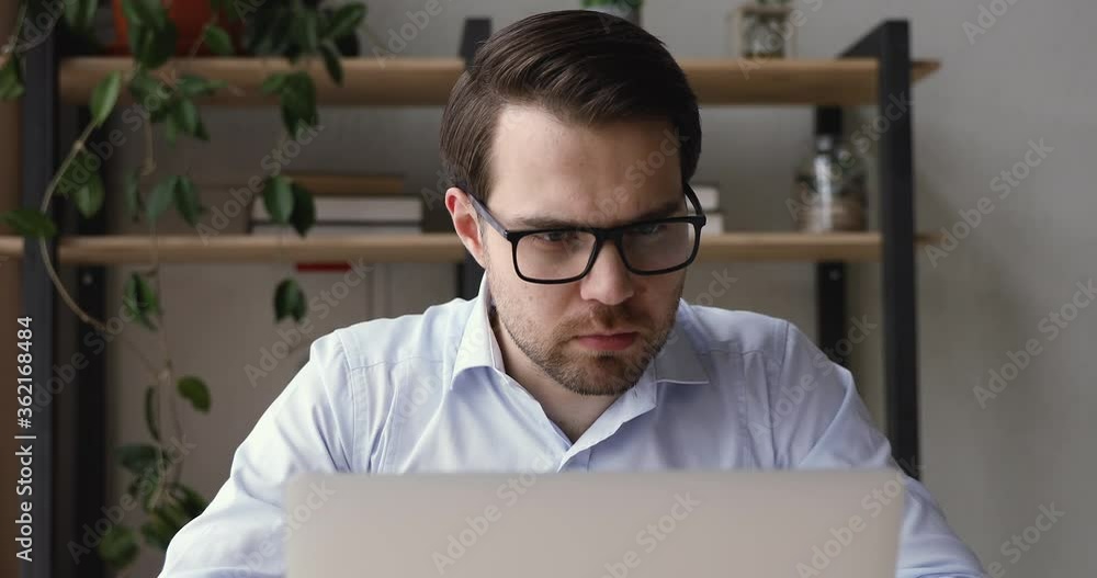 Millennial caucasian businessman sit at desk indoor wearing eyeglasses working using laptop learn new application having questions thinking search solution having fruitful busy workday in office room Millennial caucasian businessman sit at desk indoor wearing eyeglasses working using laptop learn new application having questions thinking search solution having fruitful busy workday in office room