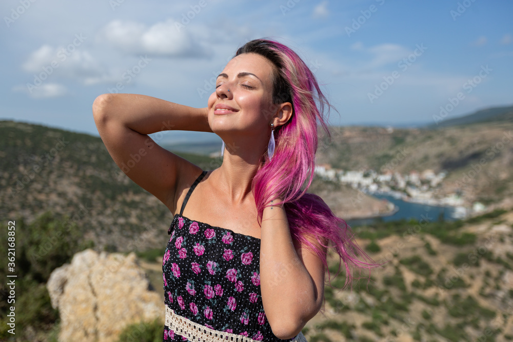A beautiful girl with rosy cheeks and pink hair, closed her eyes and enjoys the sun on the background of mountain ranges and parts of the sea A beautiful girl with rosy cheeks and pink hair, closed her eyes and enjoys the sun on the background of mountain ranges and parts of the sea
