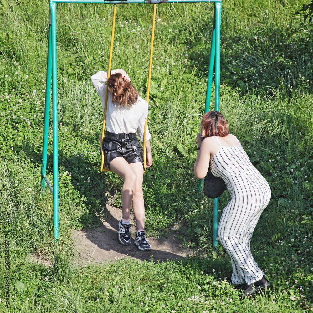 A young European girl in a short skirt poses for a photographer on a swing on a Sunny summer day in a Park against a background of green grass
