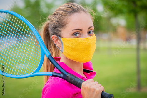 Portrait of tennis player girl holding racket outside with protective masks - Close up blonde girl who is playing tennis during quarantine holding racket in hand, preparing to play tennis outdoors 