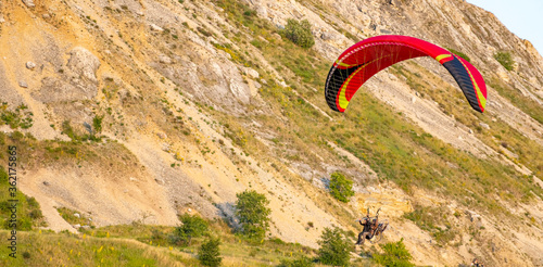 A tandem of people under the red dome of a motor paraglider takes off into the air in a mountain gorge. Red paraglider on the background of lime mountain.