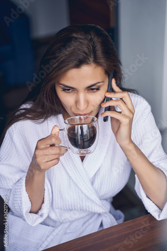Charming young woman drinking coffee and having phone conversation