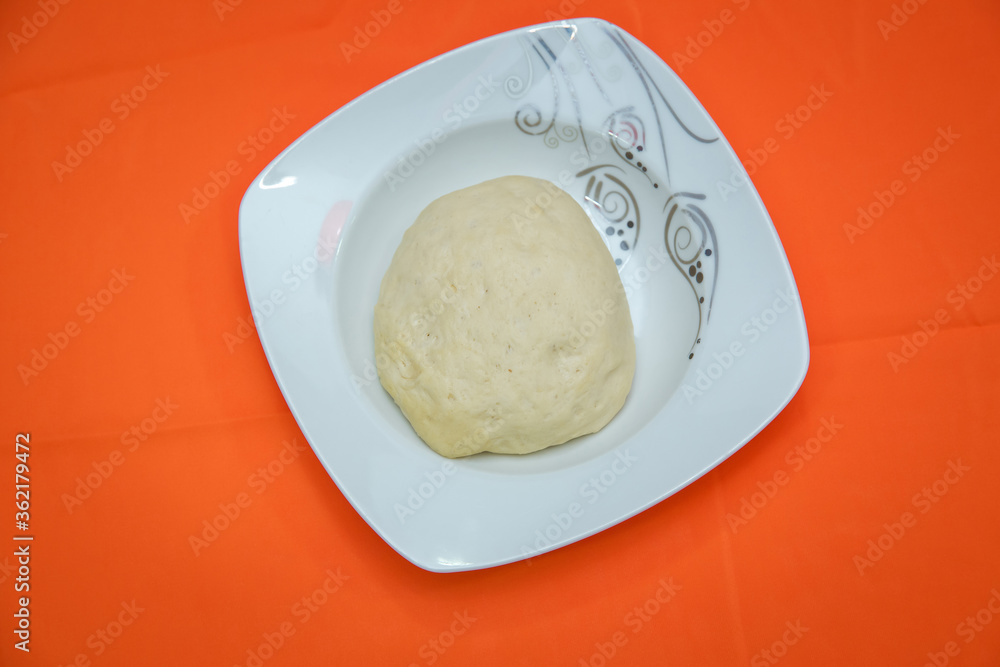 Raw dough lying on the plate on a white background. A piece of dough for bread baking . The dough is on a white plate on an orange background . Raw dough lying on the plate on a white background. A piece of dough for bread baking . The dough is on a white plate on an orange background .