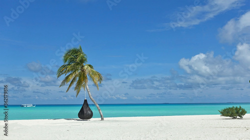 Fototapeta Naklejka Na Ścianę i Meble -  Romantic maldivian landscape. On a white sand stands a palm tree with fluttering leaves and a swing, next to a green bush. A boat floats across the turquoise ocean. Picturesque clouds on the azure sky