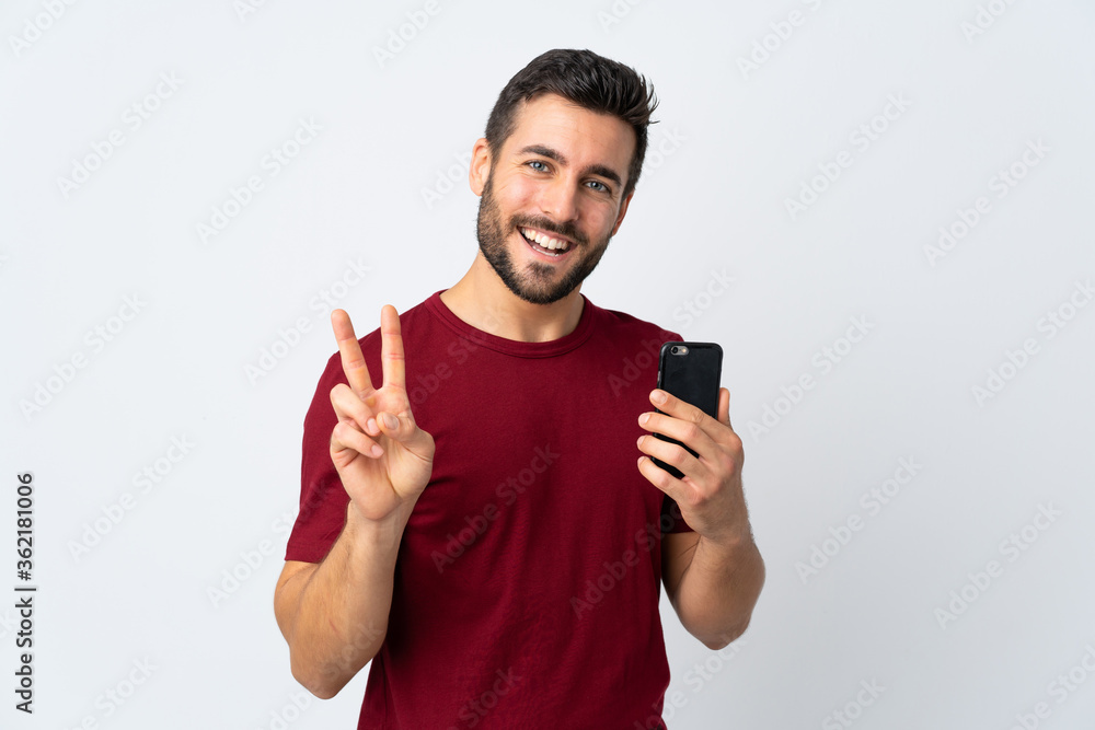 Young handsome man with beard using mobile phone isolated on white background smiling and showing victory sign Young handsome man with beard using mobile phone isolated on white background smiling and showing victory sign
