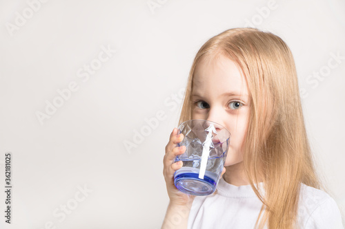 A beautiful girl of Caucasian appearance drinks water from a glass on a white background. A young blonde model girl holds a glass of water.