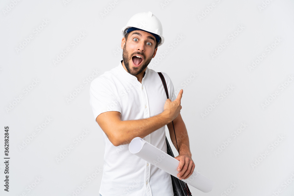 Young architect man with helmet and holding blueprints isolated on white background surprised and pointing side Young architect man with helmet and holding blueprints isolated on white background surprised and pointing side