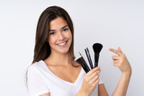 Teenager girl over isolated background holding makeup brush and pointing it
