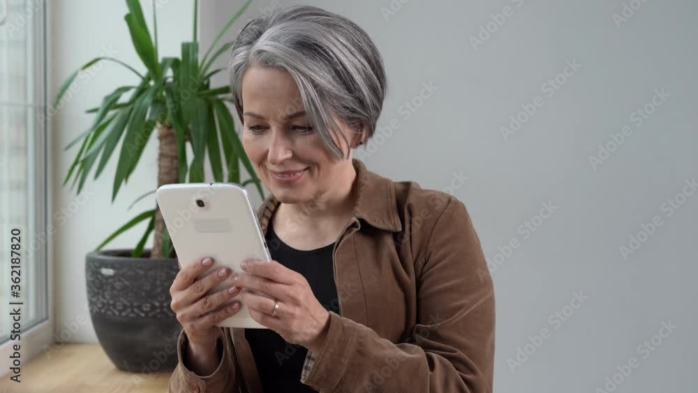 Mature woman use digital tablet lift head and smiling at camera. Mature woman in brown jacket and jeans holds tablet computer standing near window. Prores 422.