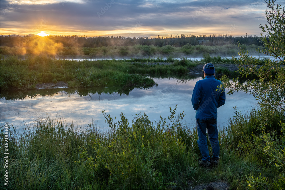 Man standing near watching the sunrise over the mountains and under clouds with reflection in the nearby river, Madison River, Montana