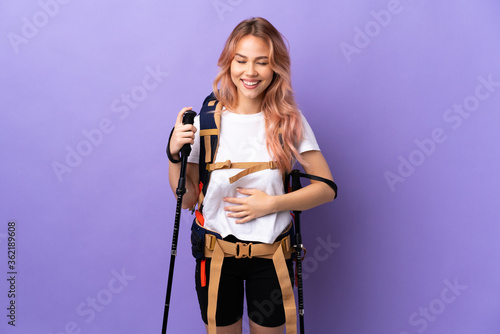 Teenager girl with backpack and trekking poles over isolated purple background smiling a lot