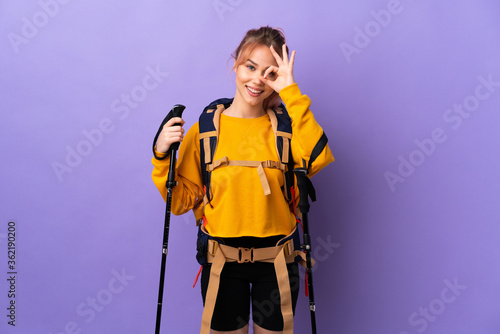 Teenager girl with backpack and trekking poles over isolated purple background showing ok sign with fingers