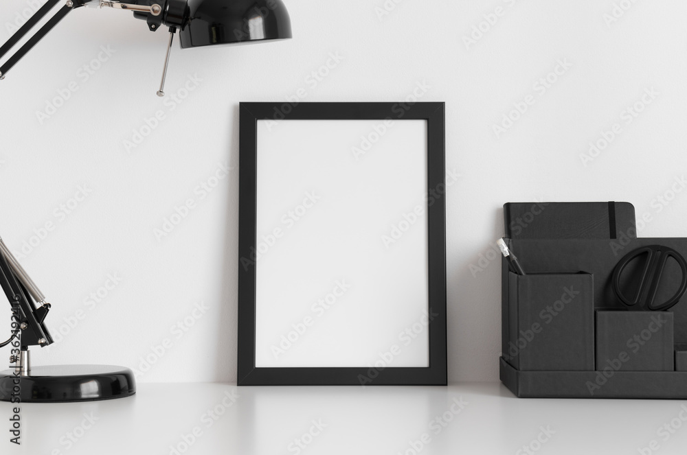 Black frame mockup with a lamp and workspace accessories on a white table. Portrait orientation.