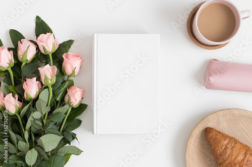 Top view of a white book mockup with a bouquet of pink roses, coffee and a croissant on a white table.