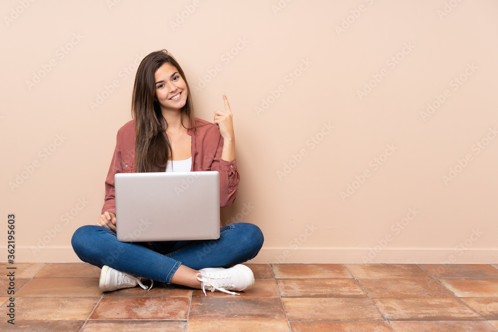 Teenager student girl sitting on the floor with a laptop showing and lifting a finger in sign of the best Teenager student girl sitting on the floor with a laptop showing and lifting a finger in sign of the best