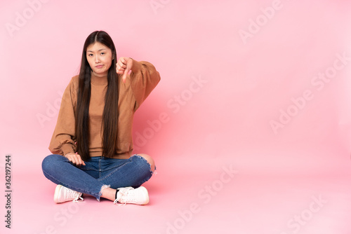 Young asian woman sitting on the floor isolated on pink background showing thumb down with negative expression