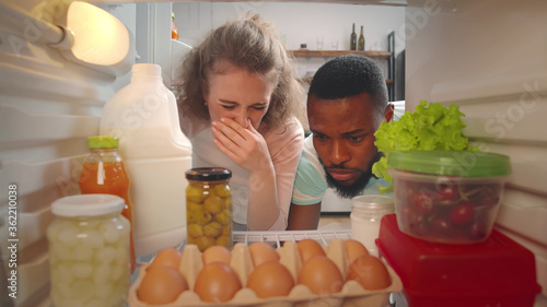 View from inside fridge of young multiracial couple feeling bad smell of spoiled meal. Woman closes her nose photo