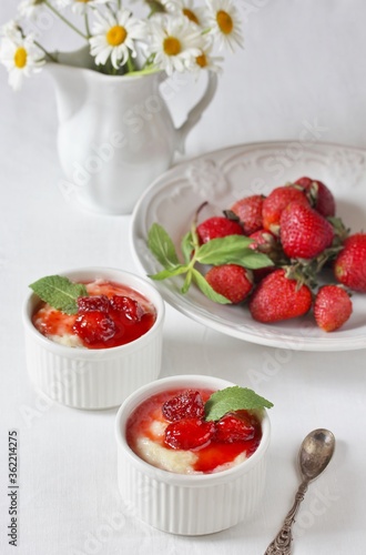 Pudding from semolina in the form of "ramekin" for baking with strawberry syrup and strawberries on a white tablecloth. daisies in a white pitcher. Soft focus