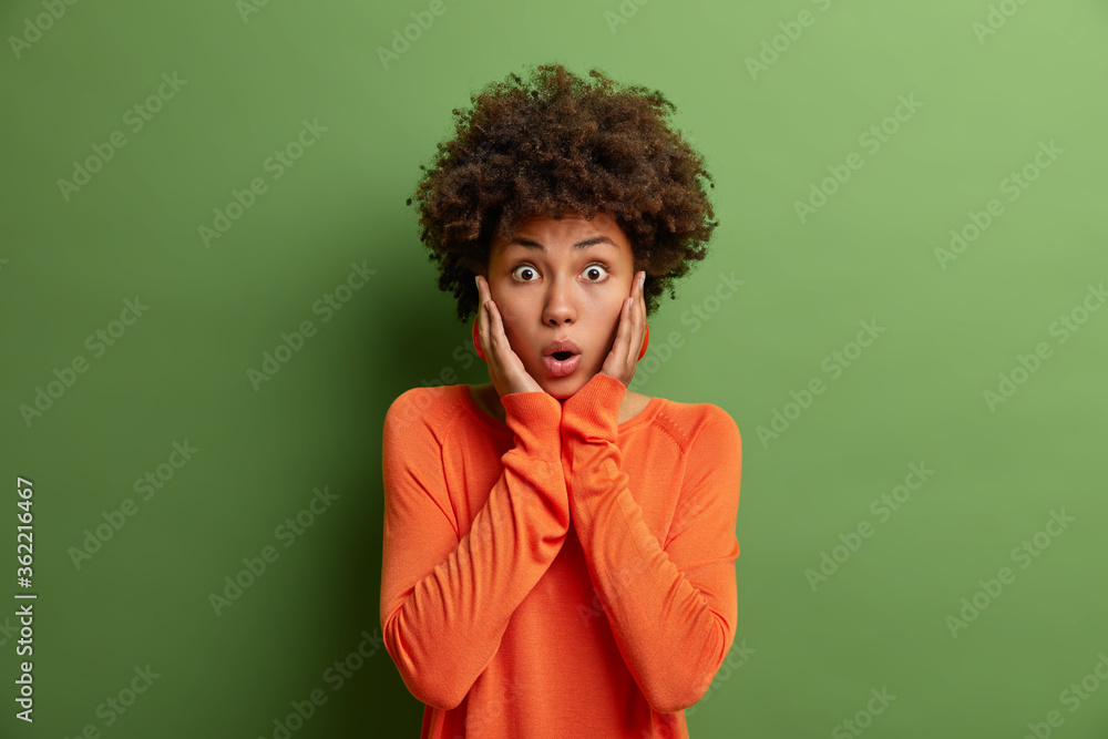 Photo of surprised astonished curly woman scared and shocked by crazy news, touches cheeks and stares at camera, holds breath in sudden fright, wears orange jumper, isolated on green background Photo of surprised astonished curly woman scared and shocked by crazy news, touches cheeks and stares at camera, holds breath in sudden fright, wears orange jumper, isolated on green background