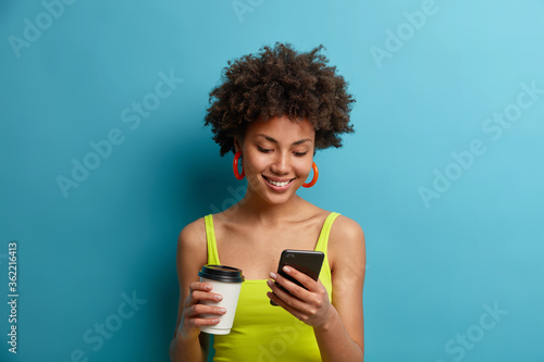 Smiling happy curly haired woman downloads new messanger or makes shopping online, drinks takeaway coffee, wears bright vest, being addicted to modern technologies, isolated on blue studio wall