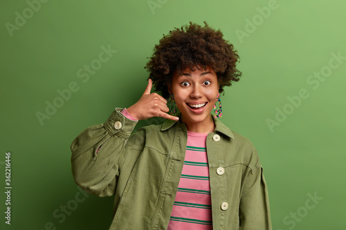 Happy positive African American woman makes call me gesture, smiles happily and asks to contact with her, has friendly expression, encourages to go in cafe together, communicates on distance