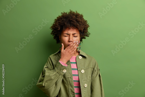 Sleepy exhausted African American woman covers mouth with palm, needs sleep, wakes up early in morning, hates Monday as to go for work, dressed in green clothes, feels very tired after party