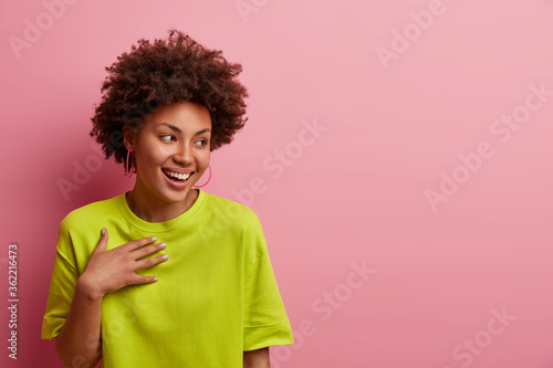 Cheerful carefree woman laughs at something positive, looks away with glad expression, cannot hide happiness after attending concert of favorite singer, poses against pink background, empty space