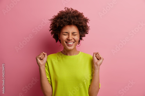 Joyful emotive dark skinned woman raises clenched fists, makes triumph gesture, cheers about success, smiles broadly, feels encouraged and motivated, looks upbeat as has great day, grabed opportunity