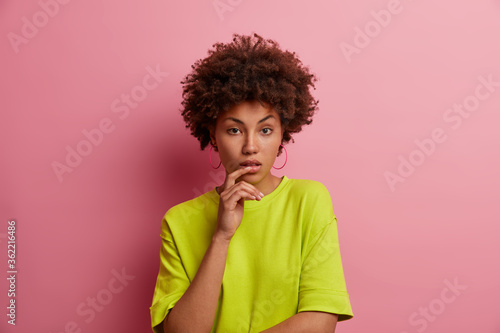 Portrat of beautiful serious unsmiling woman with calm attentive focused expression  keeps finger on lips and listens carefully  looks directly at camera  dressed in casual outfit  isolated on pink