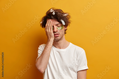 Image of exhausted student makes face palm, feels tired and overworked because of working all night, stands with drowsy expression, needs sleep, applies patches for refreshing look. People, insomnia © wayhome.studio 