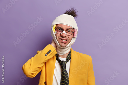 Fotografiet Displeased young man with shiner under eye, suffers from neck pain, looks dissatisfied at camera, wears bandage around injured head, becomes victim of severe fight, isolated on purple background