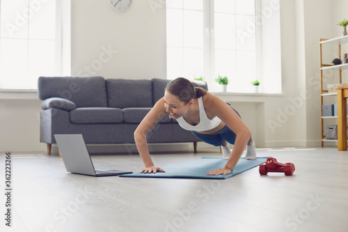 Fototapeta Naklejka Na Ścianę i Meble -  Beautiful fit girl in sportswear watching workout video online and doing push ups or plank exercise at home