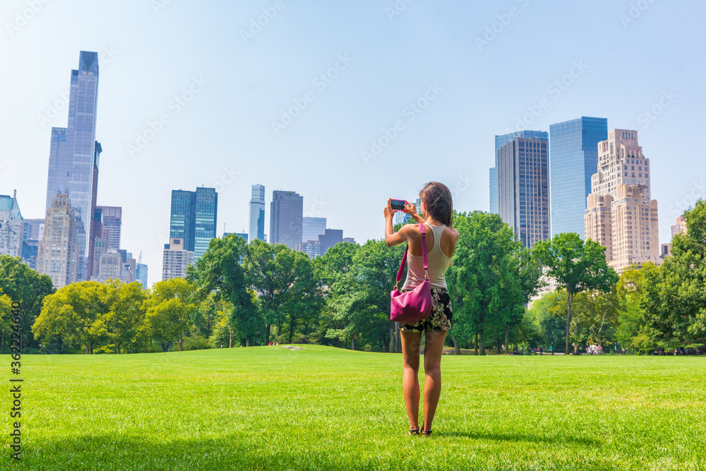 New York City woman tourist taking photo with phone of NYC Skyline in Sheep Meadow Central Park. Summer travel vacation landscape manhattan walk activity.