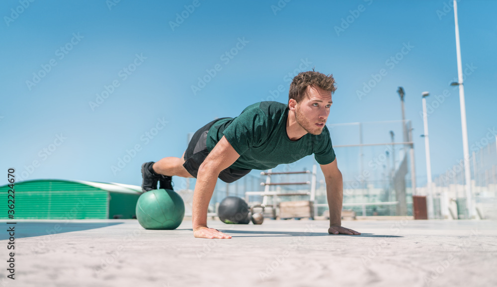 Fit exercises man strength training core doing balance push-ups workout at outdoor gym balancing on stability medicine ball with legs. Bodyweight pushups exercises. Push-up variation.