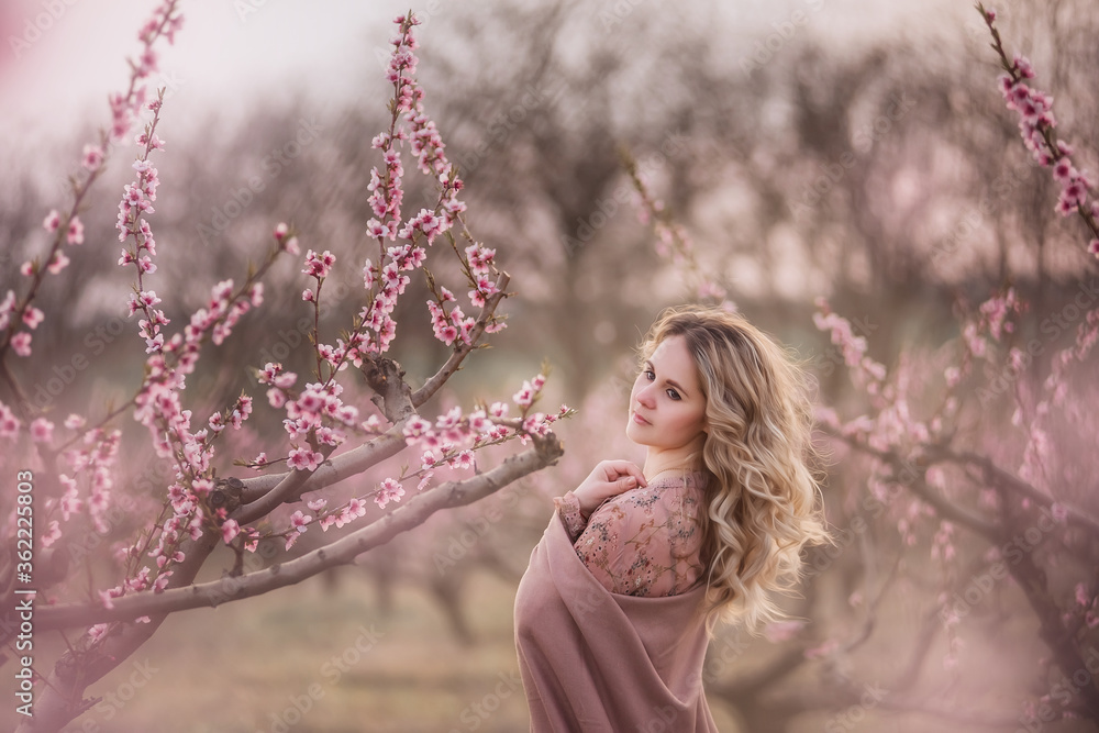 Tender fresh young girl with beautiful curly blonde hair, with a clean face, natural makeup, stands in blooming gardens, holds a twig with pink inflorescences. Mysterious, pensive, sensitive woman Tender fresh young girl with beautiful curly blonde hair, with a clean face, natural makeup, stands in blooming gardens, holds a twig with pink inflorescences. Mysterious, pensive, sensitive woman