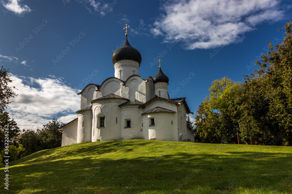 Ancient white orthodox christian stone temple. Pskov region, Russia. Church of St. Basil the Great on a hill against a background of green grass and a beautiful blue sky with white clouds. 