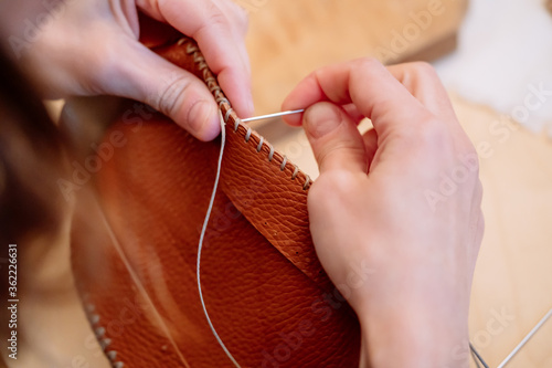 Detail shot of woman's hands sewing a brown piece of leather with a needle