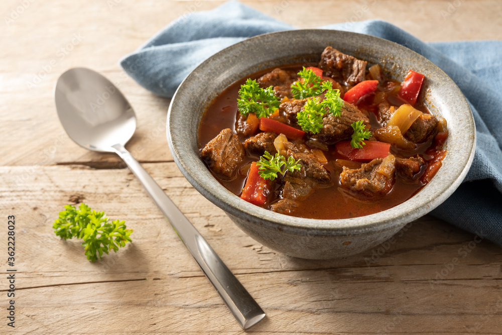 Fresh cooked goulash or stew from beef, onions and red bell pepper with parsley garnish in a bowl with spoon and a blue napkin on a rustic wooden table, Fresh cooked goulash or stew from beef, onions and red bell pepper with parsley garnish in a bowl with spoon and a blue napkin on a rustic wooden table,
