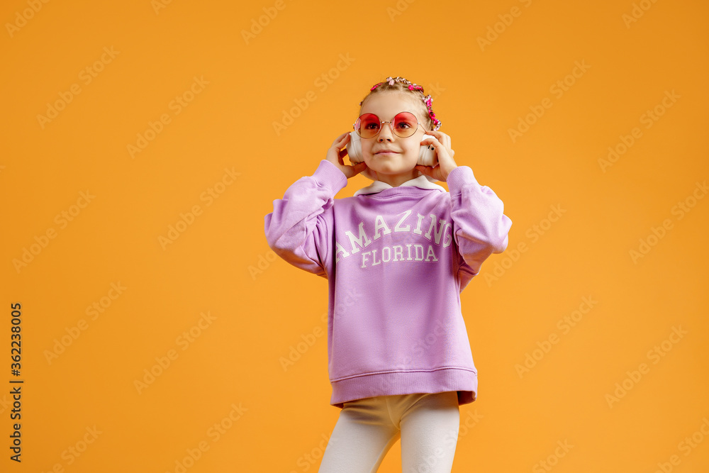 Adorable child in rounded glasses with pink dreadlocks listen to music playing in earphones on yellow background. Cute small kid listening to music in headset, copy space. Adorable child in rounded glasses with pink dreadlocks listen to music playing in earphones on yellow background. Cute small kid listening to music in headset, copy space.