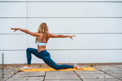 Fototapeta Naklejka Na Ścianę i Meble -  Yoga with blonde caucasian girl exercising on a yellow mat with a white wall in the background, with a blue maya on her legs and a white short shirt. On your knees with your arms open back