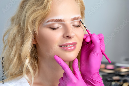 Blonde woman having permanent make-up tattoo on her eyebrows. Closeup beautician doing tattooing eyebrow. Green eyes close up. Professional makeup and cosmetology skin care.