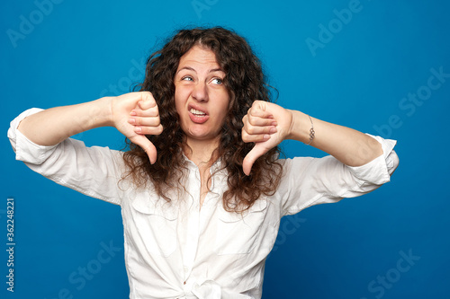 Curly haired female expresses dislike with body language sign , keeps thumbs down and frowns face, has displeased angry expression, sun-tanned skin, curves lips, isolated over blue wall background.